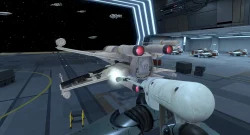 First-person view from a pilot in a white spacefighter, aiming a forward cannon inside a futuristic hangar with ships docked along the bay.