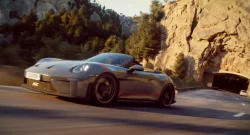 Silver Porsche sports car speeding along a mountain road with rocky cliffs and trees, approaching a tunnel on the right side of the scene.