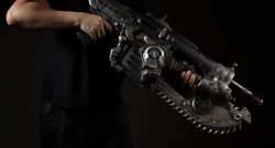 Person in a black shirt holding a large futuristic gun with a rotating chainsaw-style blade along the underside, against a dark background.