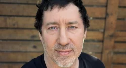 Close-up portrait of a man with short dark hair and light stubble against a wooden fence backdrop.