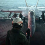 Pilot in a helmet and flight suit stands on an aircraft carrier deck beside a parked fighter jet, facing the aircraft.