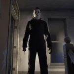 Masked killer in black coveralls stands on a dim staircase landing, holding a knife, in a dark, eerie hallway.