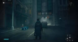 Person in a long coat seen from behind walks down a dim city street at night, with a car ahead and signs glowing in foggy light.