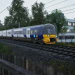 Modern passenger train with a yellow front on elevated tracks beside a gray office building, with overhead power lines and surrounding trees.